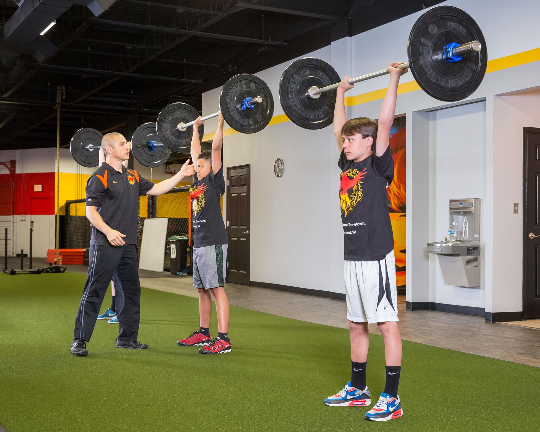Two young teen boys, each holding a barbell over their heads while their coach looks on