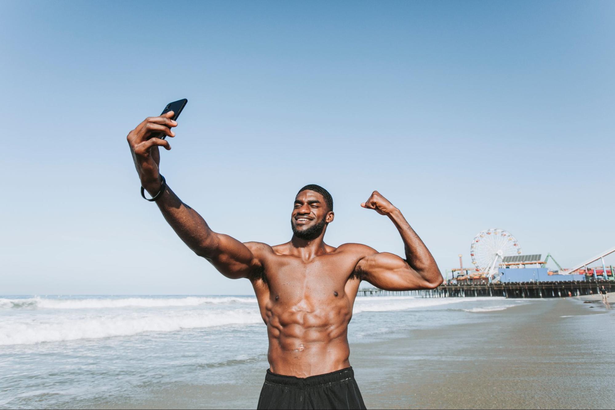 Fit man taking a selfie on the beach while flexing