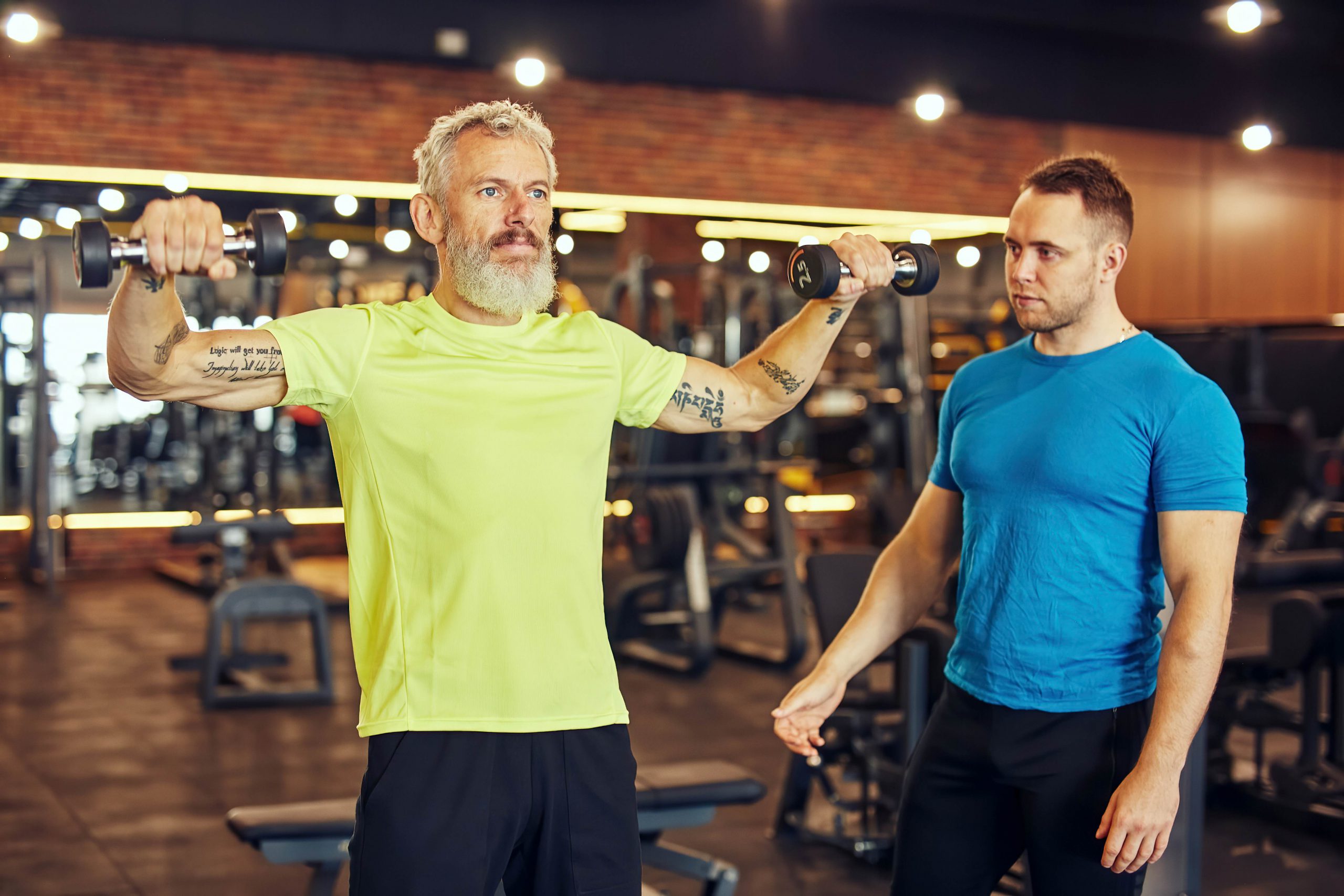 Focused mature man in sportswear exercising with dumbbells under supervision of a young professional personal trainer at gym